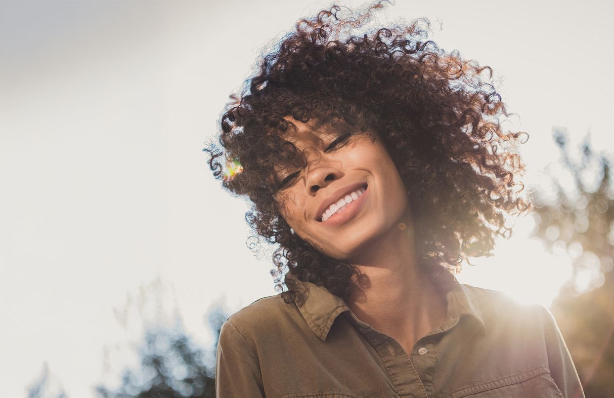 Smiling woman with curly hair, sunlight backdrop.